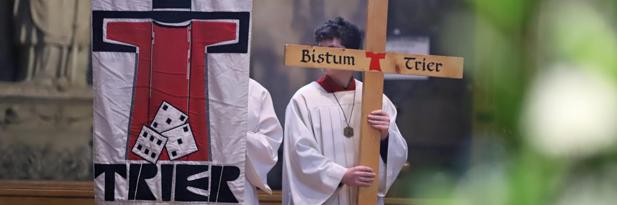 Zwei Messdiener in einer Messe; einer hält die Pilgerfahne mit der Aufschrift 'Trier' hoch, der andere ein Kreuz, auf dem Bistum Trier steht.