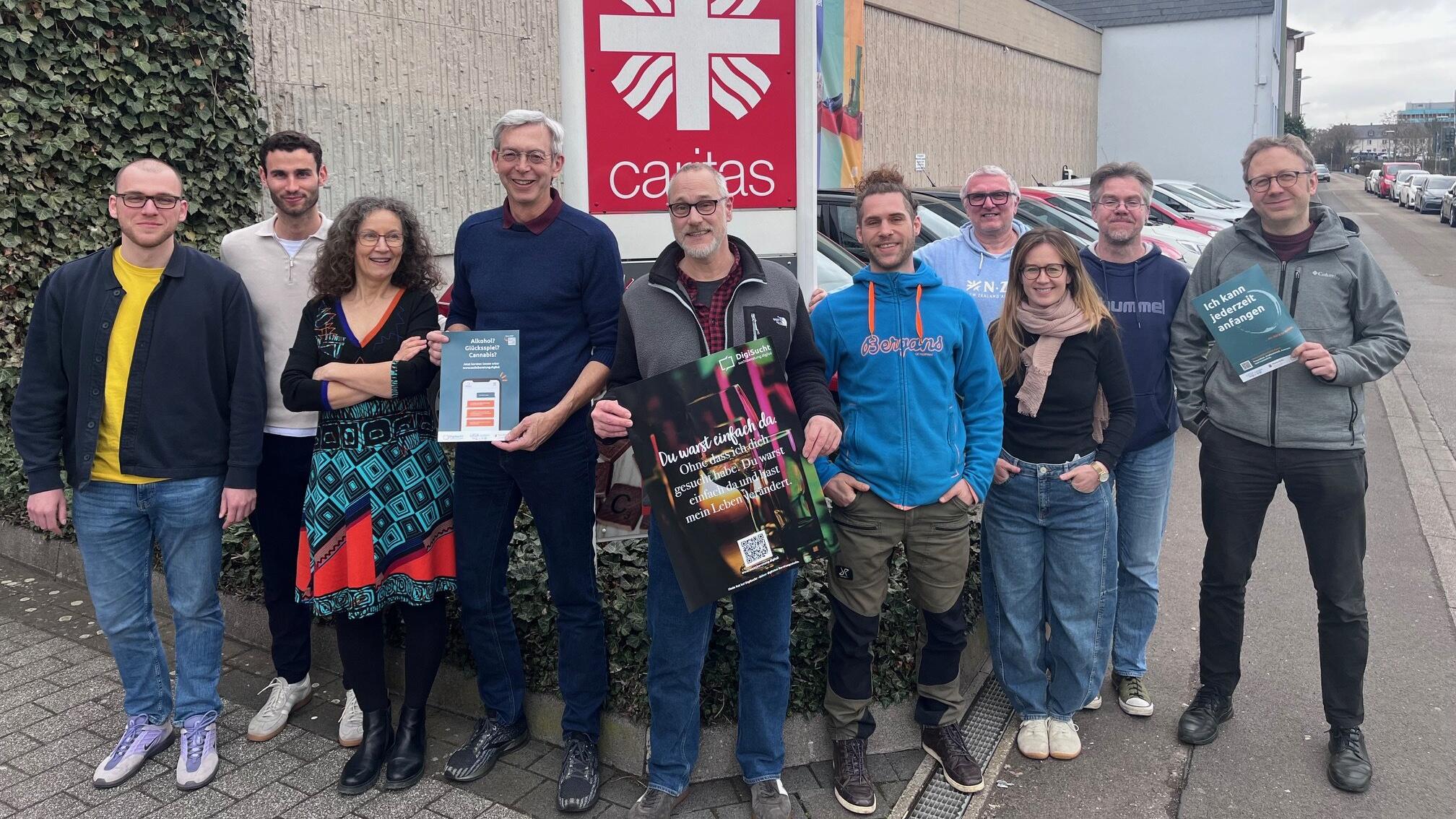 Eine Gruppe von unterschiedlichen Leuten steht draußen vor dem Caritas-Logo und schaut in die Kamera.